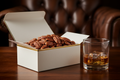 Give me a sees candy box bourbon pecans inside, sitting on a dark wooden table, there should be a leather chair in the background and a glass of bourbon next to it.